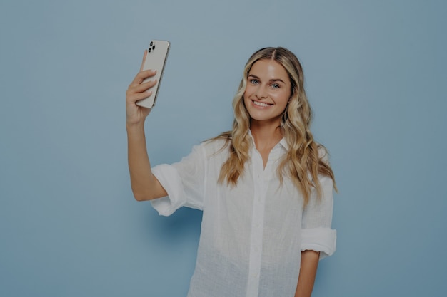 Mulher jovem sorridente e alegre com cabelo loiro comprido ondulado fazendo foto no smartphone