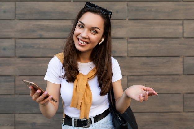 Mulher jovem sorridente e atraente, vestindo roupas casuais, em pé na rua, segurando e usando