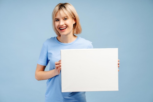 Mulher jovem sorridente e bonita com aparelhos dentários segurando um cartaz em branco para texto isolado Mockup