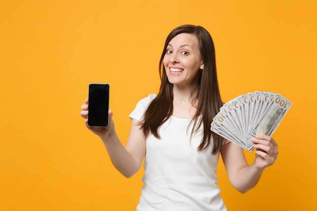 Mulher jovem sorridente em roupas brancas segura celular com ventilador de tela vazia em branco de dinheiro em notas de dólar isoladas em fundo amarelo laranja. Conceito de estilo de vida de pessoas. Mock up espaço de cópia.