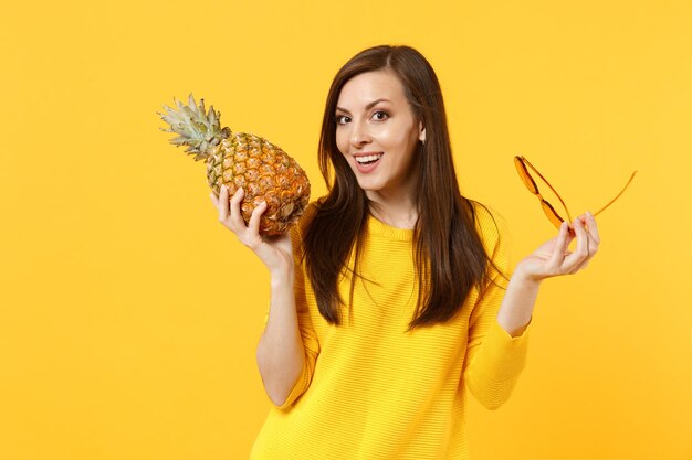 Mulher jovem sorridente em roupas casuais segura nas mãos frutas frescas de abacaxi maduro, óculos de sol isolados no fundo da parede laranja amarela. O estilo de vida vívido das pessoas relaxa o conceito de férias. Espaço de cópia de mock up