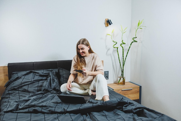 Mulher jovem sorridente em roupas casuais sorrindo e trabalhando remotamente sentada na cama com o laptop durante o trabalho remoto Trabalho freelancer Trabalhe em casa