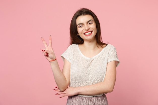 Mulher jovem sorridente em roupas leves casuais posando isolado no fundo da parede rosa pastel, retrato de estúdio. Conceito de estilo de vida de emoções sinceras de pessoas. Mock up espaço de cópia. Mostrando sinal de vitória.