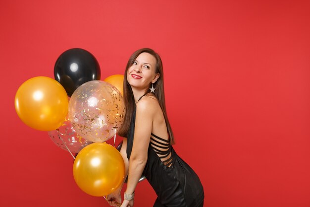 Foto mulher jovem sorridente em um vestido preto comemorando olhando para cima e segurando balões de ar isolados em fundo vermelho brilhante. dia de são valentim, feliz ano novo, conceito de festa de feriado de maquete de aniversário.