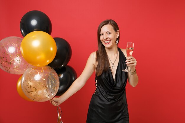 Mulher jovem sorridente em um vestido preto comemorando, segurando a taça de champanhe, balões de ar isolados sobre fundo vermelho. Dia de São Valentim, feliz ano novo, conceito de festa de feriado de maquete de aniversário.