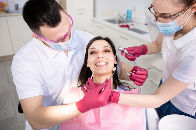 Mulher jovem sorridente em uma cadeira odontológica. Dois dentistas verificam os dentes do paciente. Viagem preventiva ao dentista. Odontologia, saúde, medicina.