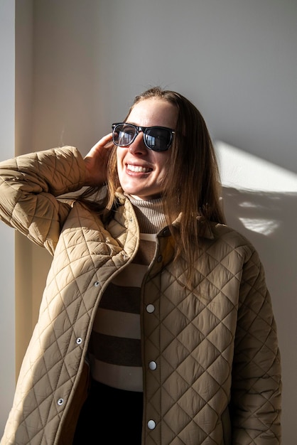 Mulher jovem sorridente em uma jaqueta em óculos de sol contra uma parede branca