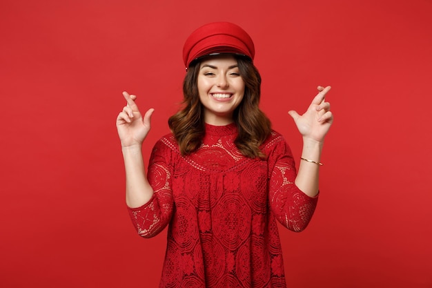 Mulher jovem sorridente em vestido de renda, boné esperando um momento especial, mantendo os dedos cruzados, fazendo desejo isolado no fundo da parede vermelha. Emoções sinceras de pessoas, conceito de estilo de vida. Mock up espaço de cópia.