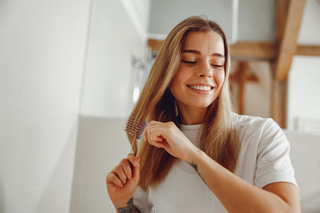 Mulher jovem sorridente escovando o cabelo com um pente enquanto está de pé no banheiro perto do espelho