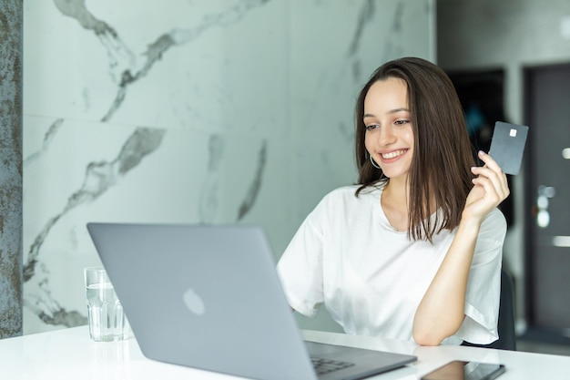 Mulher jovem sorridente fazendo compras online usando tablet e cartão de crédito na cozinha