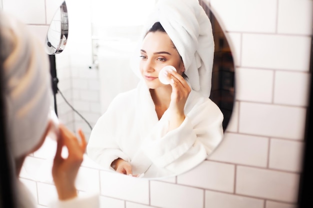 Mulher jovem sorridente lavando o rosto com esponja de limpeza facial no banheiro.