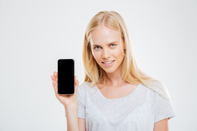 Mulher jovem sorridente mostrando a tela do smartphone em branco, isolada em uma parede branca