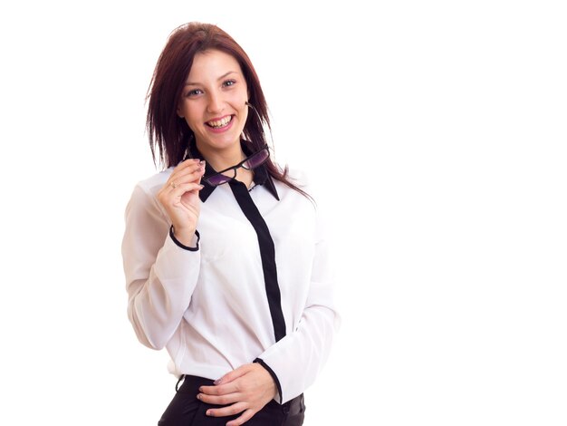 Mulher jovem sorridente na camisa branca e calça preta com cabelo escuro segurando óculos escuros em fundo branco no estúdio