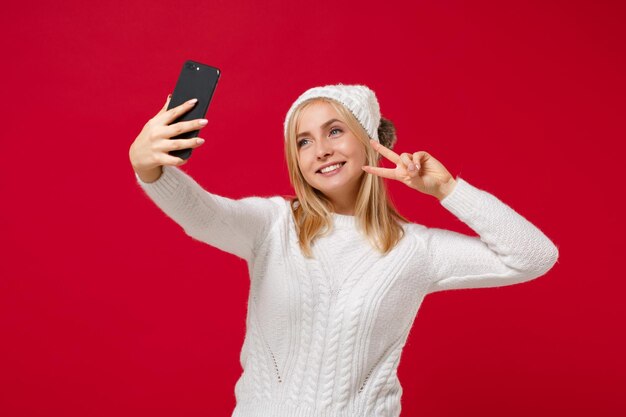 Mulher jovem sorridente na camisola branca, chapéu isolado no fundo da parede vermelha. Estilo de vida de moda saudável, conceito de estação fria. Simule o espaço da cópia. Fazendo um tiro de selfie no celular, mostrando sinal de vitória.