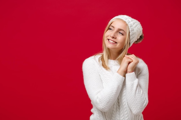 Foto mulher jovem sorridente na camisola branca, chapéu isolado no fundo da parede vermelha, retrato de estúdio. estilo de vida de moda saudável, conceito de estação fria. simule o espaço da cópia. olhando de lado, segurando as mãos cruzadas.