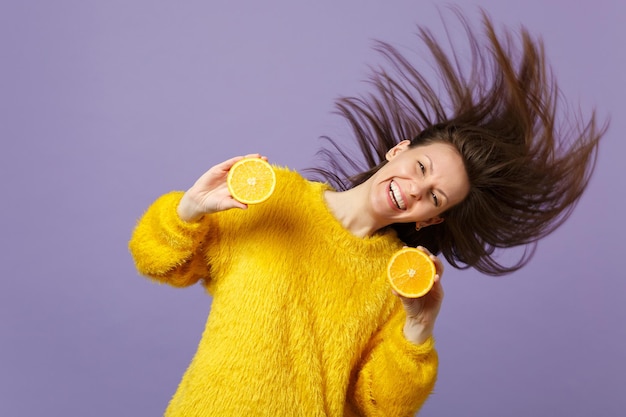 Mulher jovem sorridente no suéter de pele com cabelo voando segurando metades de frutas frescas laranja maduras isoladas em fundo pastel violeta. Estilo de vida vívido de pessoas, relaxe o conceito de férias. Mock up espaço de cópia.