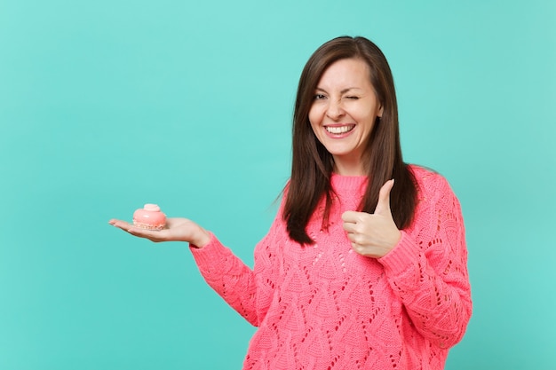 Mulher jovem sorridente no suéter rosa de malha piscando, aparecendo o polegar, segurando na mão o bolo isolado no retrato de estúdio de fundo de parede azul turquesa. Conceito de estilo de vida de pessoas. Simule o espaço da cópia.