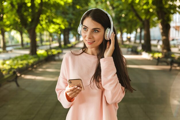Mulher jovem sorridente ouvindo música com fones de ouvido enquanto caminhava no parque da cidade segurando um telefone celular