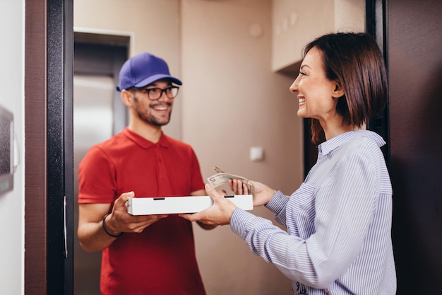 Foto mulher jovem sorridente recebendo pizza do entregador em casa.