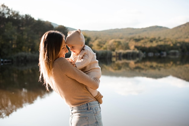 Mulher jovem sorridente segurando brincando com bebê de 1 ano de idade usa roupas de malha sobre fundo natural e lago com floresta Temporada de outono Maternidade