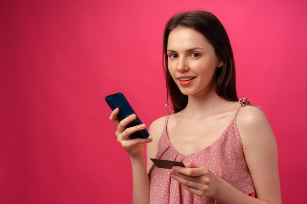 Mulher jovem sorridente segurando smartphone e cartão de crédito contra fundo rosa