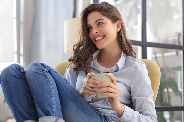 Mulher jovem sorridente sentada na poltrona da sala de estar, segurando uma xícara de café.