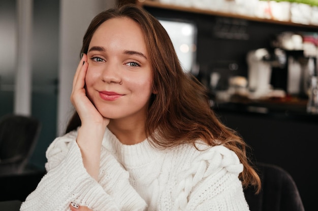 Mulher jovem sorridente sentada no café dentro de casa Garota de suéter olhando para a câmera