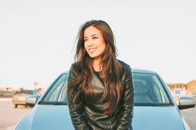 Mulher jovem sorridente sentada no capô do carro contra o céu claro
