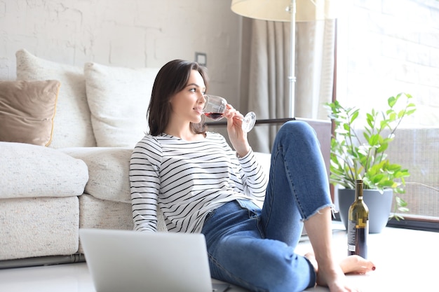 Mulher jovem sorridente, sentada no chão com o computador laptop e conversando com amigos, bebendo vinho.