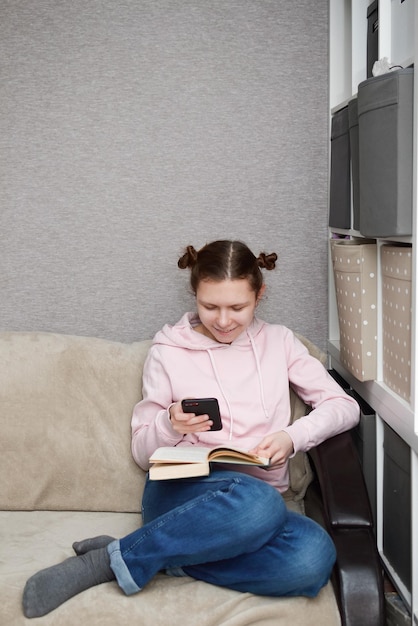 Mulher jovem sorridente sentada no sofá lendo um livro interessante distraído por um telefone celular