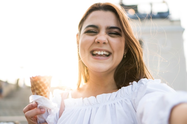 Mulher jovem sorridente tirando uma selfie enquanto toma sorvete com um pôr do sol no fundo