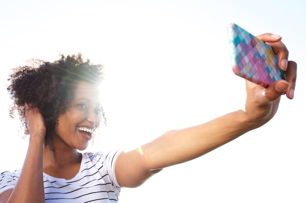 Mulher jovem sorridente tomando selfie com telefone inteligente ao ar livre contra a luz solar