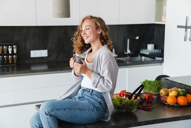 Mulher jovem sorridente, vestindo roupas casuais, tomando uma xícara de café enquanto está sentada na mesa da cozinha