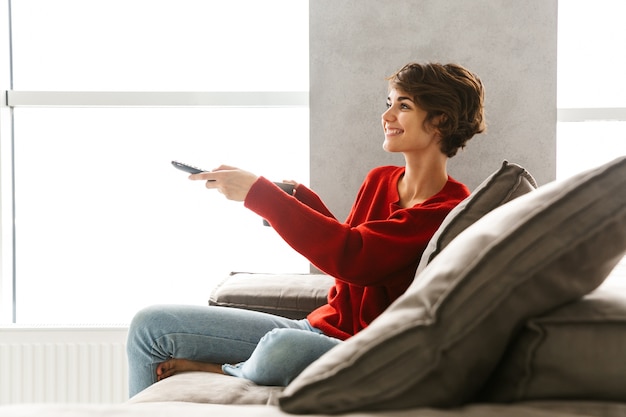 Mulher jovem sorridente, vestindo um suéter, relaxando no sofá em casa, bebendo café