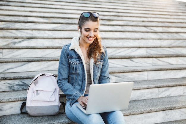 Mulher jovem sorridente, vestindo uma jaqueta, sentada na escada ao ar livre, usando um laptop