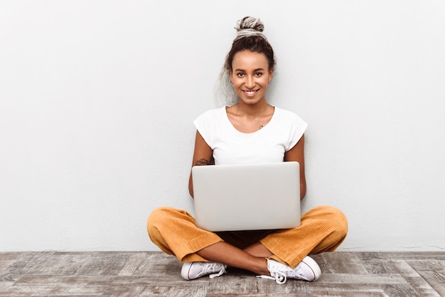 Mulher jovem sorridente, vestindo uma roupa casual, sentada isolada no branco, segurando o laptop no colo