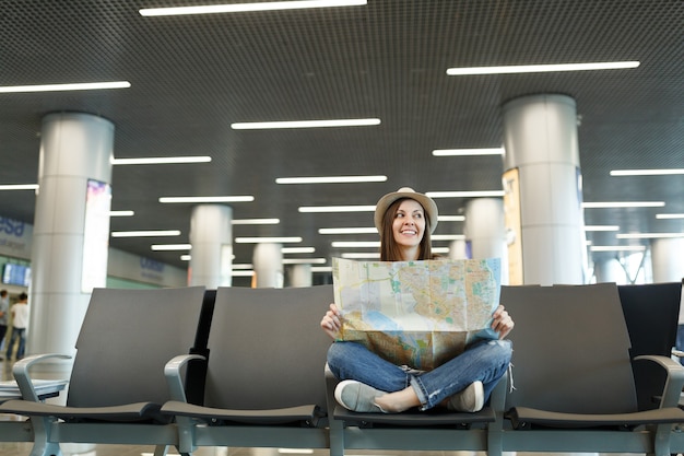 Foto mulher jovem sorridente viajante turista com as pernas cruzadas segurando um mapa de papel olhando de lado, esperando no saguão do aeroporto internacional