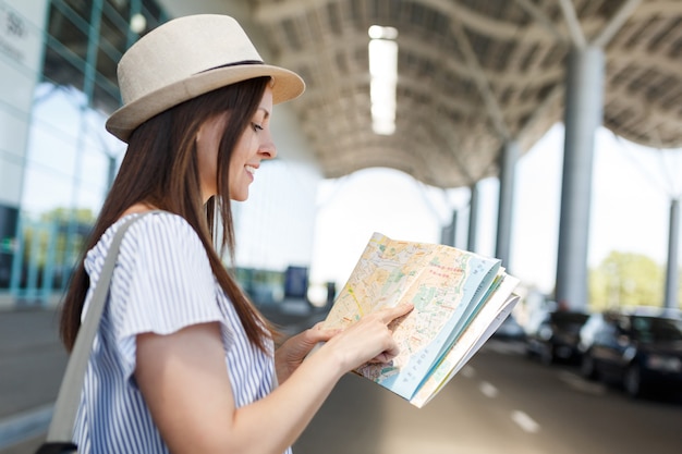 Mulher jovem sorridente viajante turista com chapéu com mochila em busca de rota no mapa de papel no aeroporto internacional