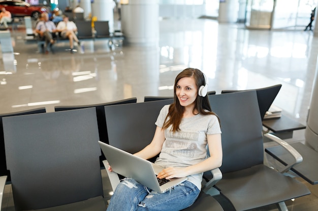 Mulher jovem sorridente viajante turista com fones de ouvido, ouvindo música trabalhando em um laptop, espere no saguão do aeroporto internacional