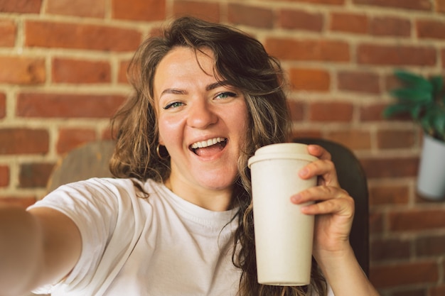 Mulher jovem sorrindo, bebendo café, olhando para a câmera e fazendo uma selfie conceito de estilo de vida