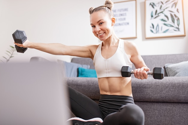 Mulher jovem sorrindo e levantando halteres enquanto assiste a um curso de fitness no laptop durante o isolamento em casa