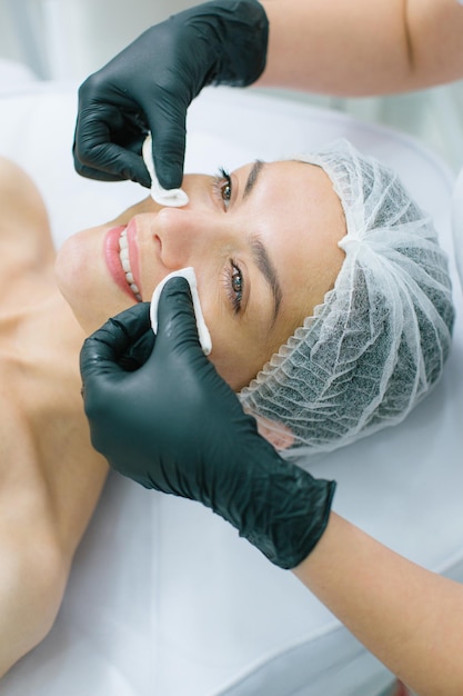 Foto mulher jovem sorrindo enquanto as mãos de um cosmetologista em luvas de borracha pretas tocam suas bochechas e as limpam