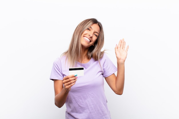 Mulher jovem sorrindo feliz e alegremente, acenando com a mão, dando as boas-vindas e cumprimentando você ou se despedindo com um cartão de crédito