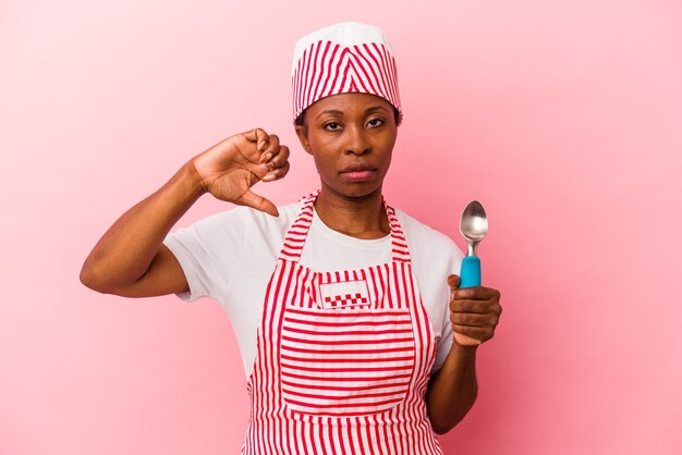 Mulher jovem sorveteira afro-americana segurando a colher isolada no fundo rosa, mostrando um gesto de antipatia, polegares para baixo. Conceito de desacordo.