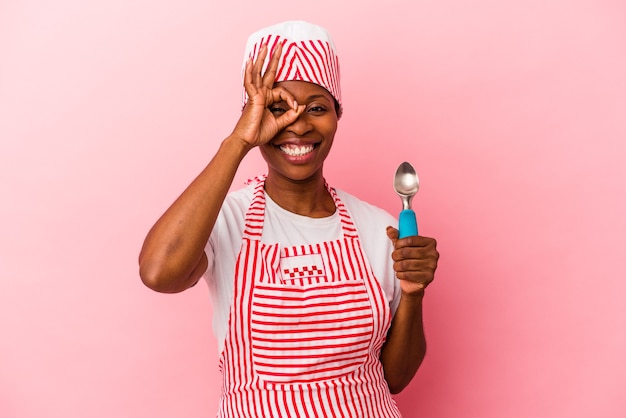 Mulher jovem sorveteira afro-americana segurando colher isolada no fundo rosa animado mantendo o gesto ok no olho.
