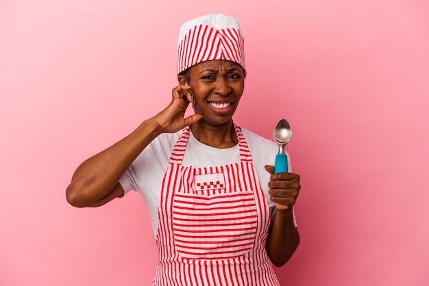 Mulher jovem sorveteira afro-americana segurando colher isolada no fundo rosa, cobrindo as orelhas com as mãos.