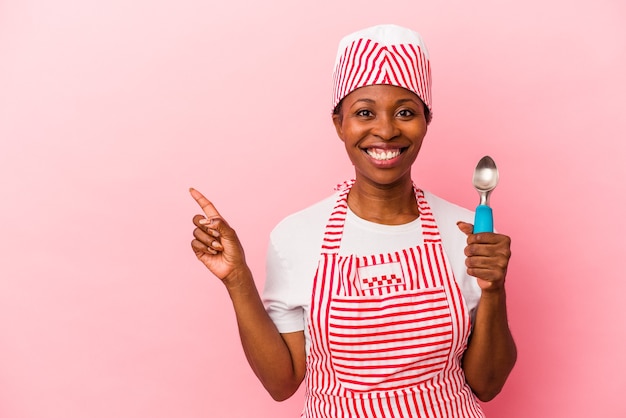 Mulher jovem sorveteira afro-americana segurando colher isolada no fundo rosa, sorrindo e apontando de lado, mostrando algo no espaço em branco.