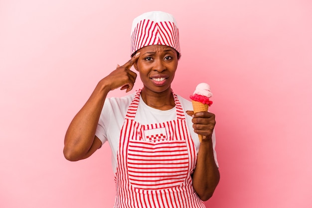 Mulher jovem sorveteira afro-americana segurando sorvete isolado no fundo rosa, mostrando um gesto de decepção com o dedo indicador.