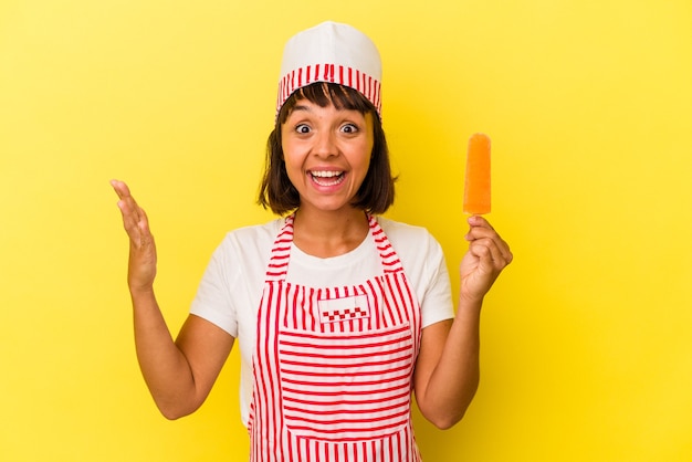 Mulher jovem sorveteira de raça mista segurando um sorvete isolado em um fundo amarelo, recebendo uma agradável surpresa, animada e levantando as mãos.