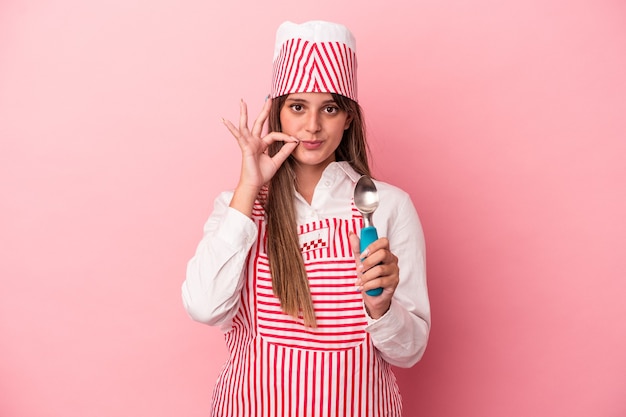 Mulher jovem sorveteira segurando uma colher isolada no fundo rosa com os dedos nos lábios, mantendo um segredo.